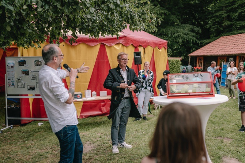 Zwei Redner stehen vor einem Publikum während einer Veranstaltung im Freien mit einem Zelt im Hintergrund.