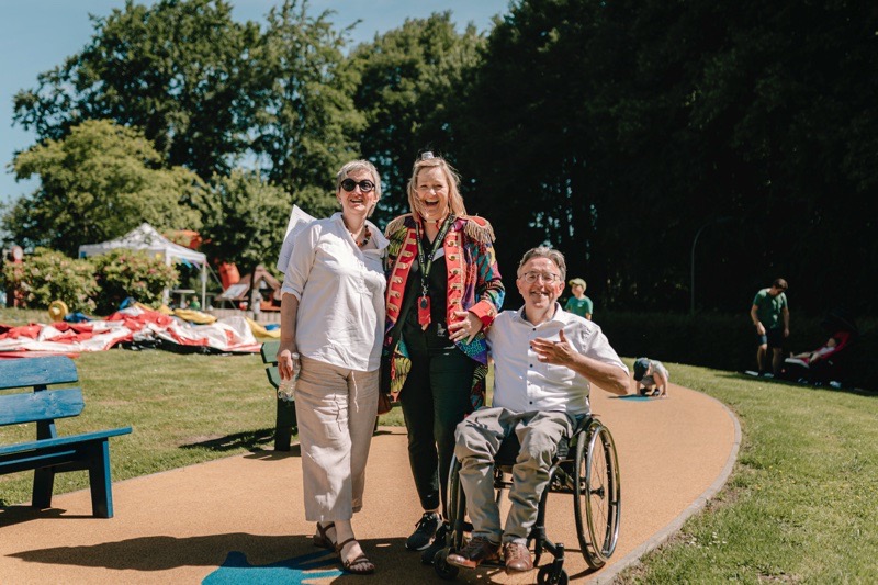 Drei lächelnde Personen stehen auf einem Weg in einem Garten, eine Person davon sitzt im Rollstuhl.