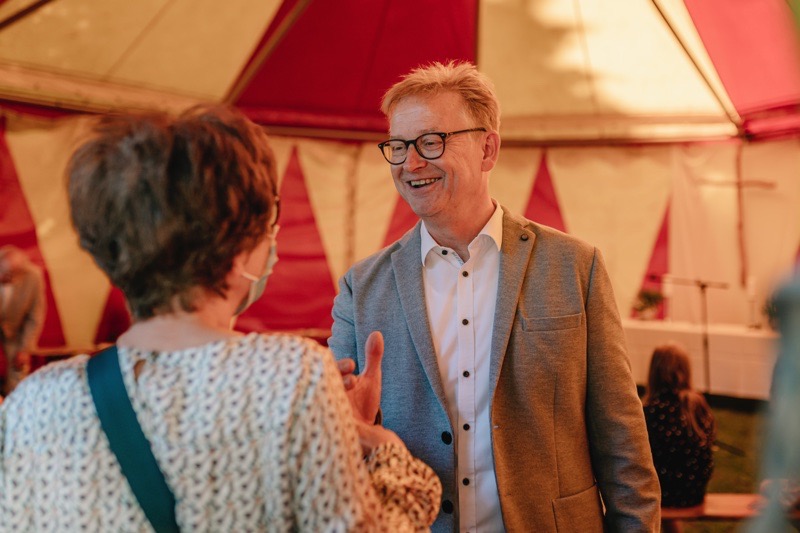 Mann mit Brille und grauem Blazer spricht mit einer Frau in einem Zelt.