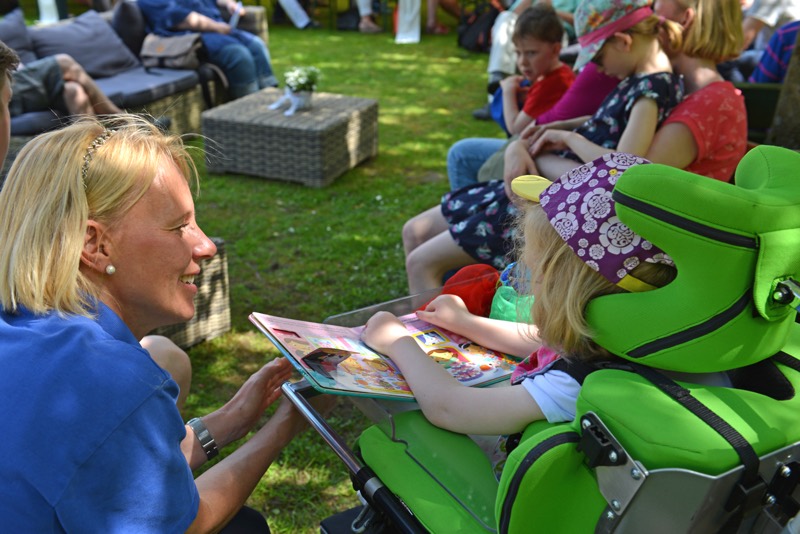 Ein Kind im Rollstuhl zeigt ein Bilderbuch einer Betreuerin während einer Veranstaltung im Freien.
