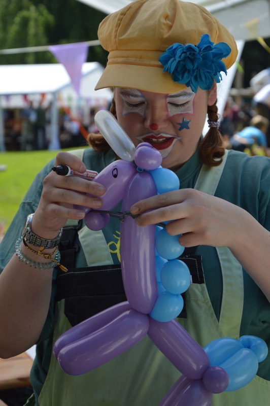 Person mit Clown-Makeup, die eine lila und blaue Ballonfigur eines Einhorns formt.