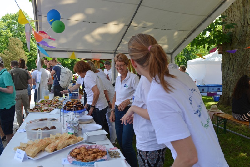 Menschen in weißen T-Shirts stehen an einem Tisch mit verschiedenen Speisen und Getränken auf einem Sommerfest.