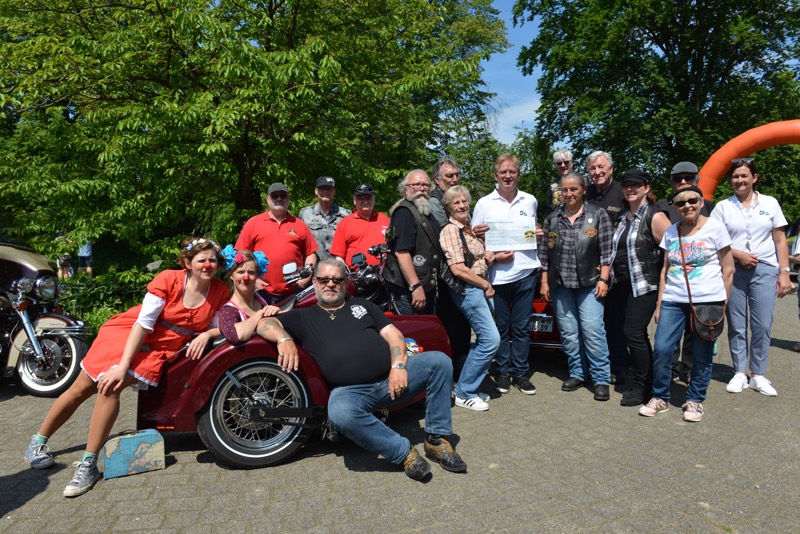 Eine Gruppe von Menschen posiert vor einem Motorrad bei einem Sommerfest im Freien.