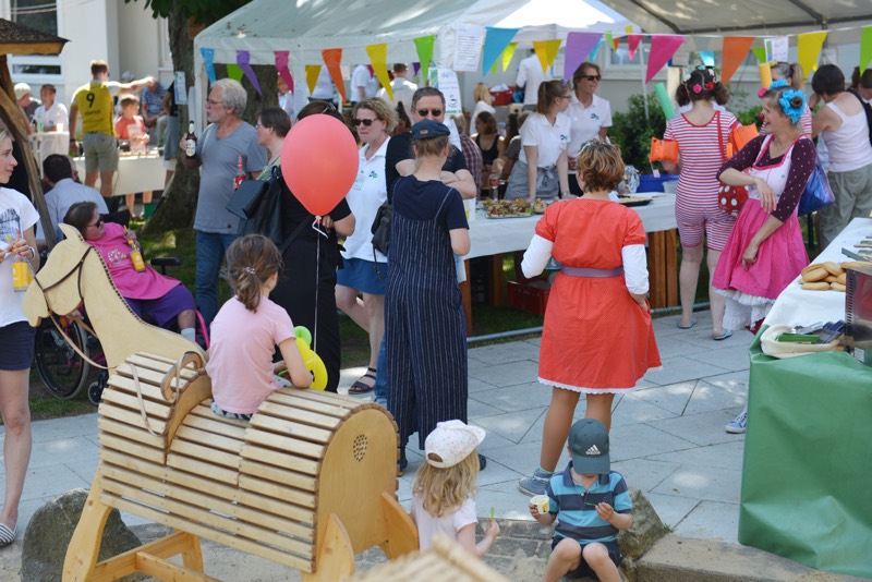 Menschen in verschiedenen Kostümen auf einem Sommerfest mit bunten Dekorationen und einem Holzspielzeugpferd.