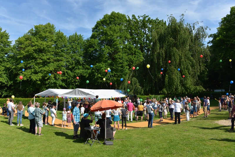 Menschenmenge beim Sommerfest lässt bunte Luftballons steigen. Zelt im Hintergrund.