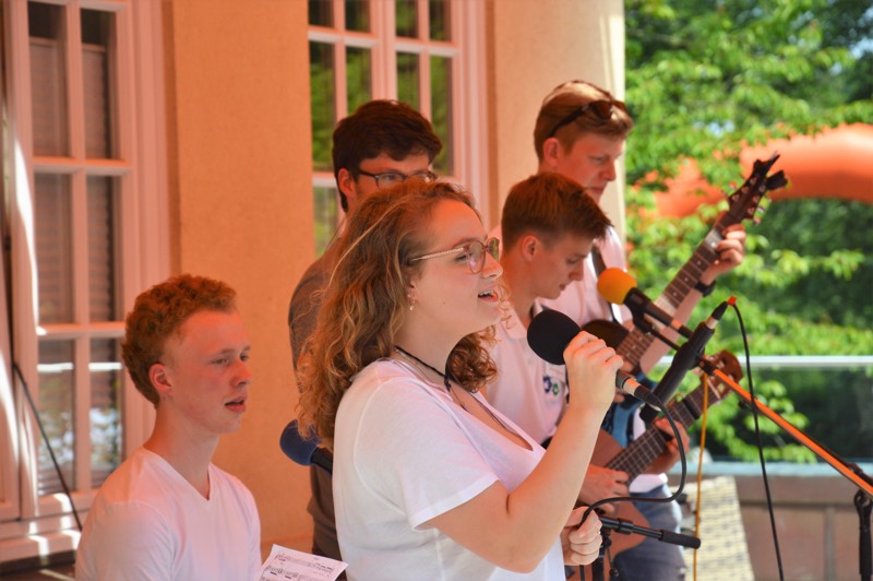 Gruppe von fünf jungen Menschen, die auf einer Bühne singen und musizieren, mit Mikrofon und Gitarren.
