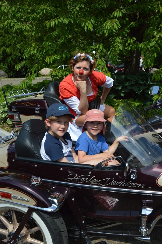 Zwei Kinder sitzen in einem Motorrad-Beiwagen, während ein Clown hinter ihnen posiert.