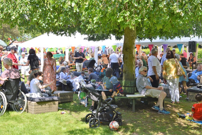 Menschen in einem Park während eines Sommerfestes, umgeben von Bäumen und bunten Fahnen.