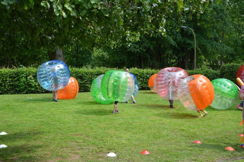 Mehrere Kinder, die in großen, transparenten Zorb-Bällen auf einer Wiese spielen.