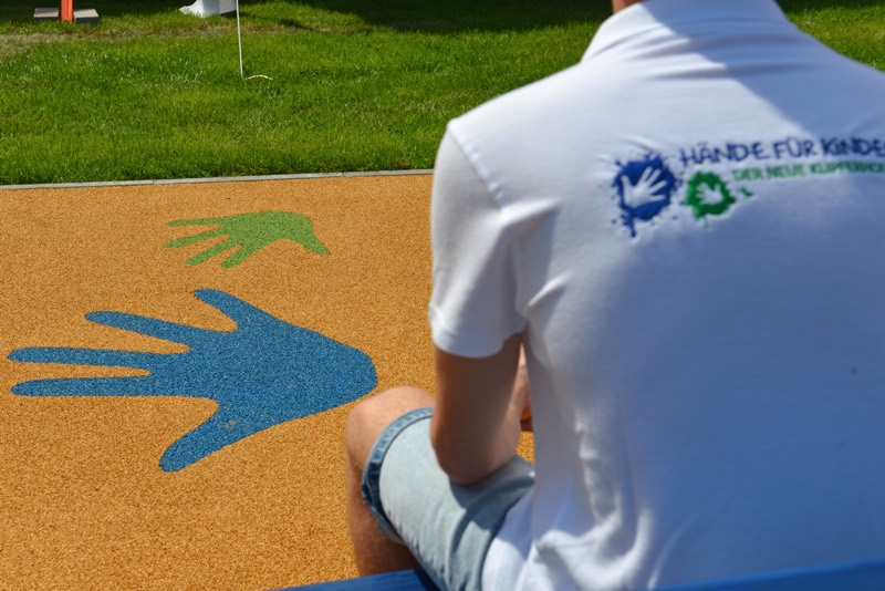 Person mit einem weißen T-Shirt, auf dem das Logo von Hände für Kinder zu sehen ist, vor einem bunten Handabdruck auf dem Boden.