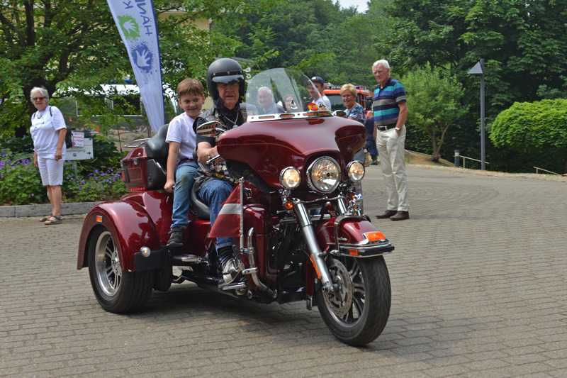 Ein rotes Dreirad-Motorrad mit einem Fahrer und einem Kind als Beifahrer auf einem Weg.