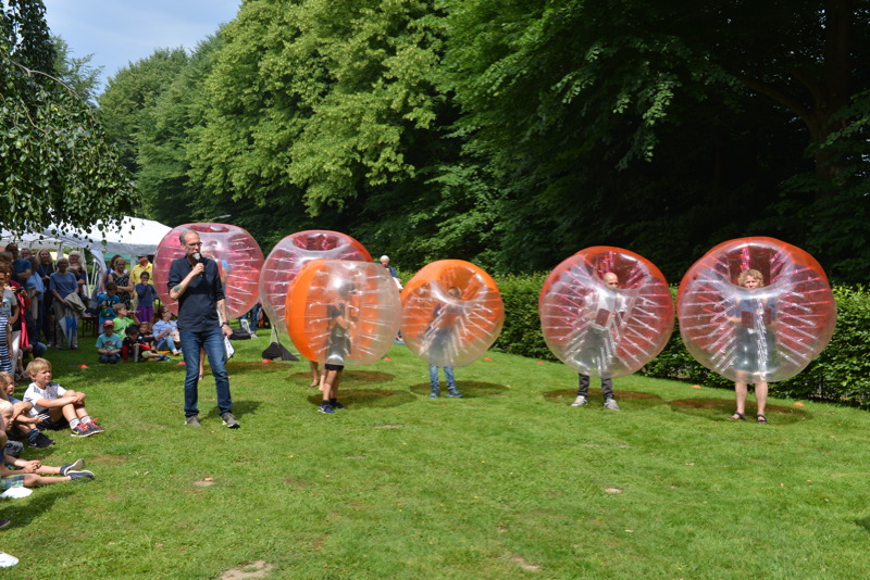 Mehrere Personen in transparenten, aufblasbaren Bällen stehen auf einer Wiese während eines Sommerfestes. Ein Mann mit Mikrofon in der Hand steht bei ihnen.