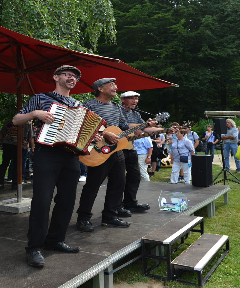 Drei Musiker auf einer Bühne, einer mit Akkordeon, zwei mit Gitarre, während sie vor Publikum spielen.