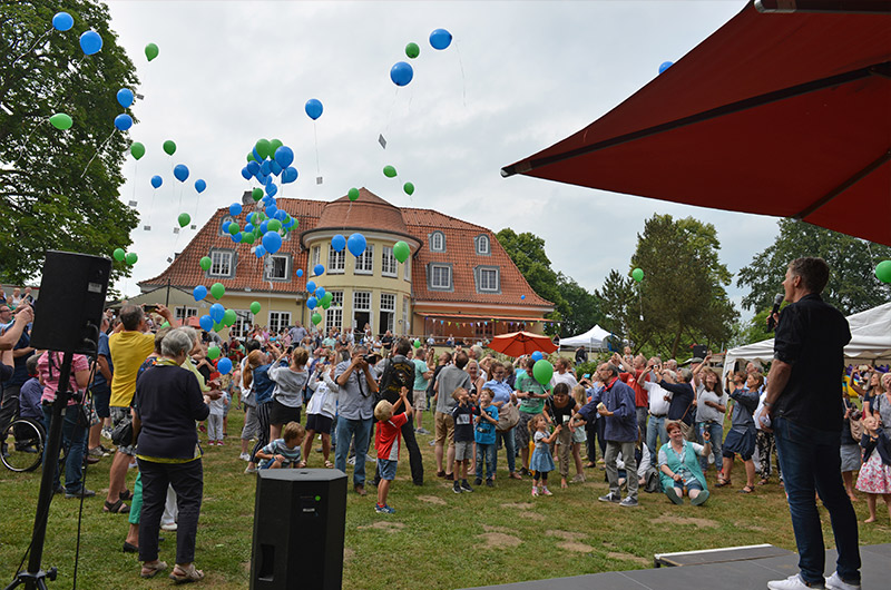 Menschenmenge, die Luftballons in verschiedenen Farben während eines Sommerfestes in einem Garten freilässt.