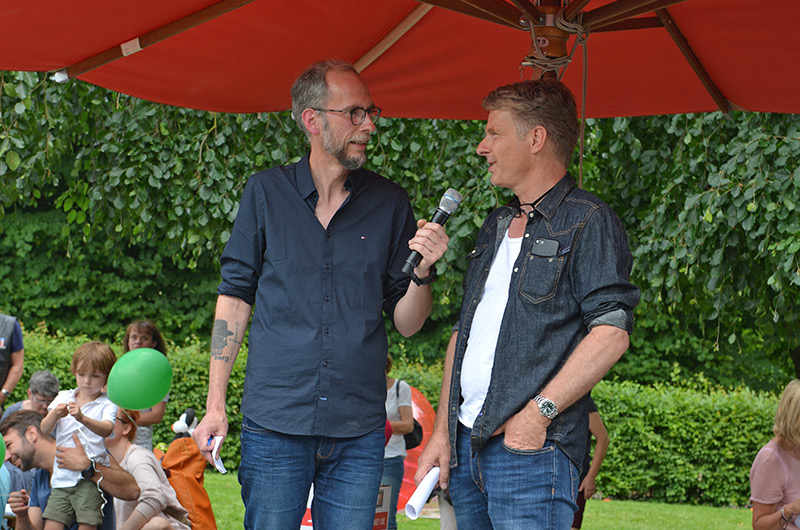 Zwei Männer stehen unter einem roten Sonnenschirm und sprechen miteinander, einer hält ein Mikrofon.