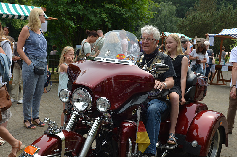 Ein Mann mit grauen Haaren fährt ein rotes Motorrad während ein Kind hinter ihm sitzt. Im Hintergrund sind viele Menschen zu sehen.