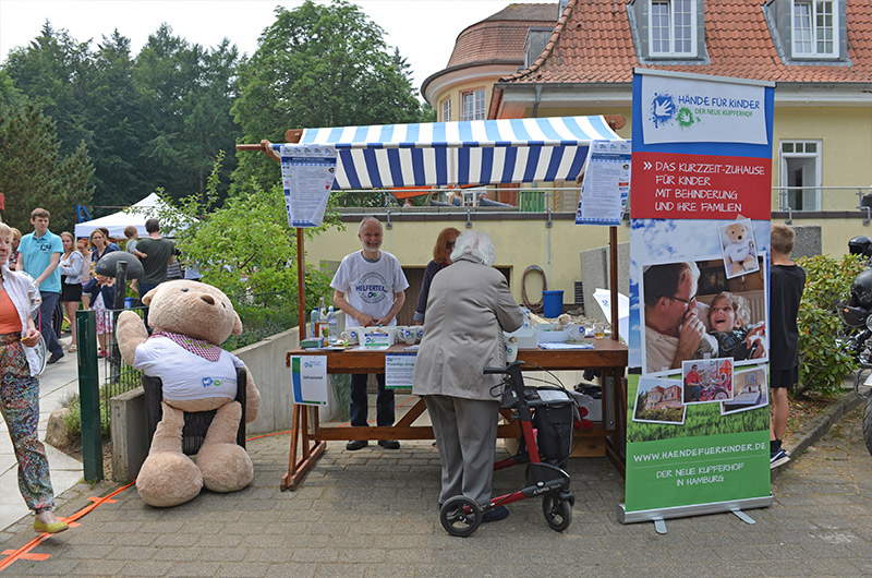 Ein Stand mit einem großen Plüschbären und einer Person, die an einem Tisch steht, bei einem Sommerfest.