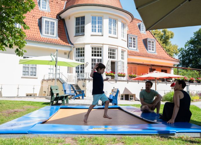 Ein Kind springt auf einem Trampolin vor dem Neuen Kupferhof, während zwei Erwachsene daneben sitzen.