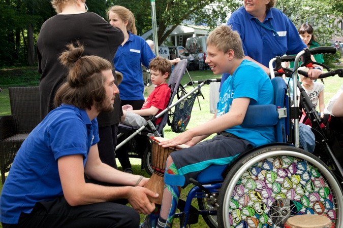 Junge im Rollstuhl interagiert mit einem Mann, der eine Trommel hält, während andere Personen im Hintergrund stehen.