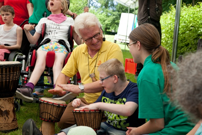 Ein Mann interagiert mit zwei Kindern, während sie auf Trommeln spielen. Eine Gruppe von Menschen ist im Hintergrund sichtbar.