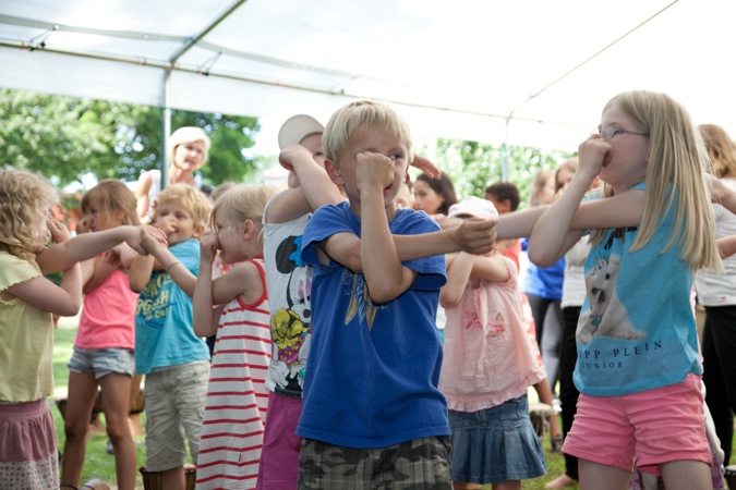 Eine Gruppe von Kindern, die an einer gemeinsamen Aktivität unter einem Zelt teilnehmen und ihre Arme bewegen.