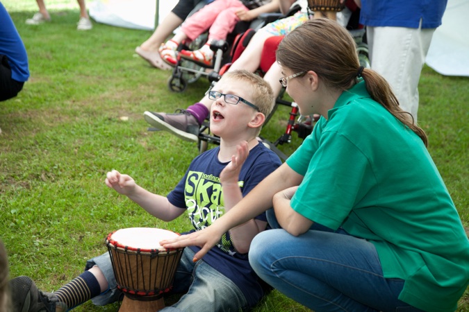 Ein Junge mit Brille spielt Trommel, während eine Betreuerin ihn unterstützt.