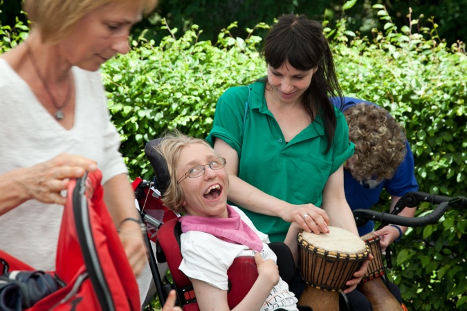Ein Mädchen im Rollstuhl lächelt, während eine Betreuerin neben ihr eine Trommel spielt.