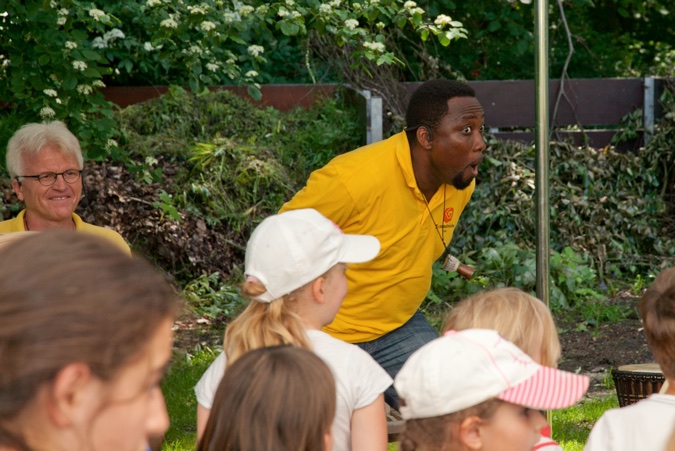 Ein Mann in gelbem Shirt führt eine Musikveranstaltung für Kinder im Freien durch.