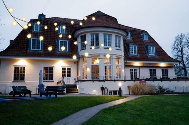 Ein großes, hell erleuchtetes Gebäude mit runden Fenstern und einer auffälligen Dachform, umgeben von Lichtern.