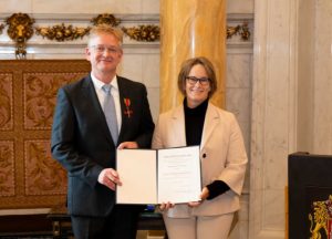 Zwei Personen stehen in einem Raum und halten eine Urkunde während einer Zeremonie.