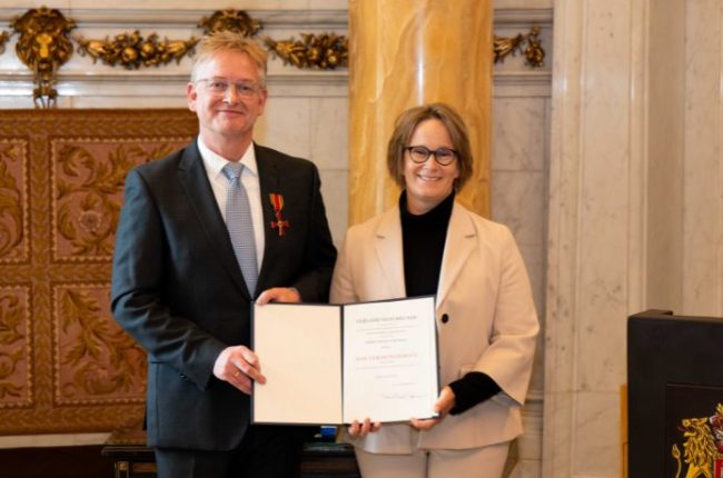 Zwei Personen stehen in einem Raum und halten eine Urkunde während einer Zeremonie.