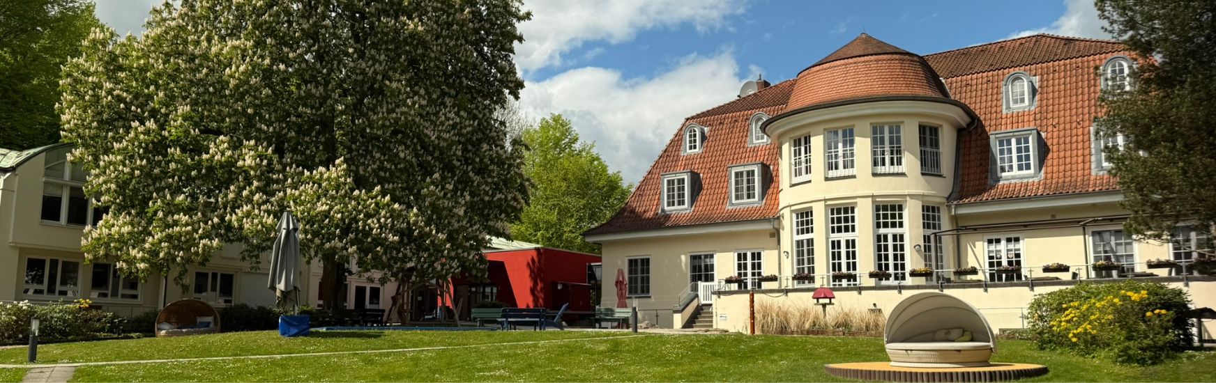 Historisches Gebäude mit runder Fassade und rotem Dach, umgeben von Bäumen und einer gepflegten Rasenfläche.