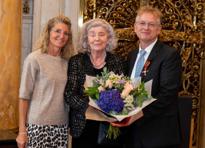 Drei Personen stehen zusammen und halten einen großen Blumenstrauß. Eine ältere Frau in der Mitte und lächelt.
