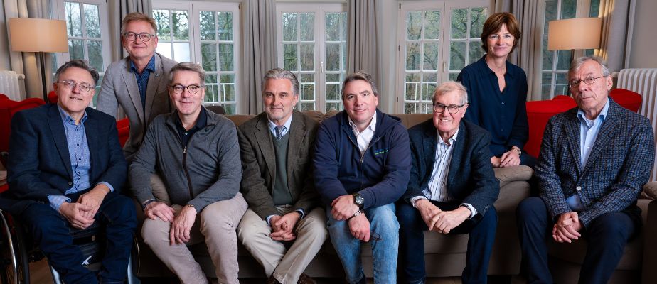 Gruppenbild von acht Personen, die in einem Raum mit Fenstern und Sofas sitzen, alle in formeller Kleidung.