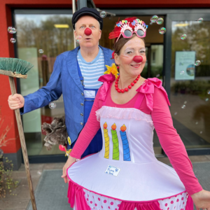 Zwei Clowns in bunten Kostümen mit roten Nasen, einer mit Besen und der andere in einem Kleid mit Kerzenmotiv.