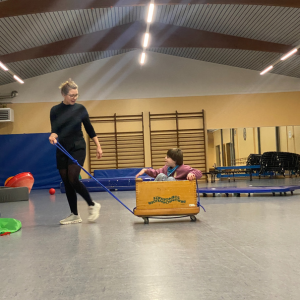 Eine Person zieht einen Kasten mit einem Kind darin durch einen Raum mit Sportgeräten und Matten.
