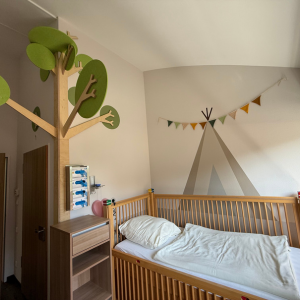 Ein Kinderbett aus Holz mit heller Bettwäsche in einem Raum mit einem Baum aus Holz und einer Zeltdekoration an der Wand.
