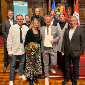 Gruppe von Personen in formeller Kleidung, einige halten Blumen, andere ein Dokument, vor einem historischen Hintergrund mit Flaggen.