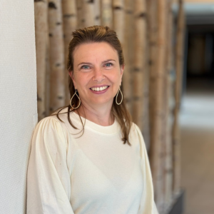 Frau mit langen, braunen Haaren und großen Ohrringen, die an einer Wand steht und lächelt.