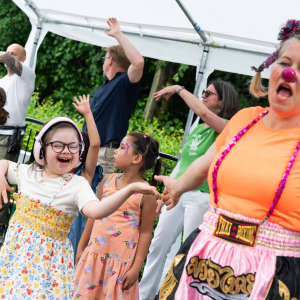 Eine Gruppe von Kindern und Erwachsenen, die fröhlich tanzen und sich bewegen, während eine Person mit Clownsnase und buntem Kostüm im Vordergrund lacht.