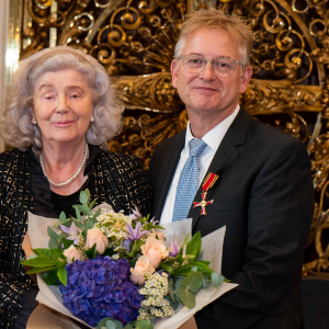 Ein Mann in einem Anzug hält einen Blumenstrauß, während eine Frau neben ihm steht. Beide posieren vor einer kunstvollen goldenen Wand.