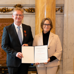 Zwei Personen stehen in einem feierlichen Rahmen und halten ein Zertifikat. Eine Person trägt eine Auszeichnung, die andere hat eine elegante Blazerjacke an.