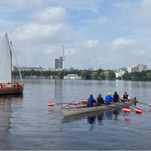 Ruderboot mit mehreren Personen auf einem ruhigen Gewässer, während ein Segelboot in der Nähe fährt.