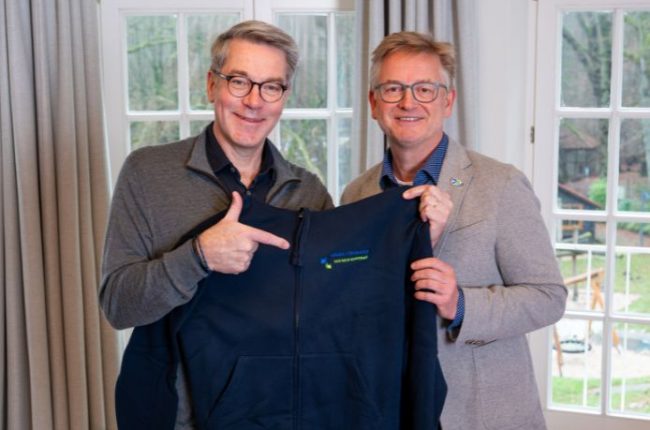 Zwei Männer posieren mit einem dunkelblauen Kapuzenpullover, der ein Logo auf der Brust zeigt. Beide lächeln in die Kamera.