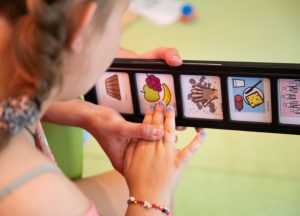 Ein Kind benutzt eine Kommunikationsleiste mit Symbolen, um sich auszudrücken. Die Symbole zeigen verschiedene Bilder wie Essen und Aktivitäten.