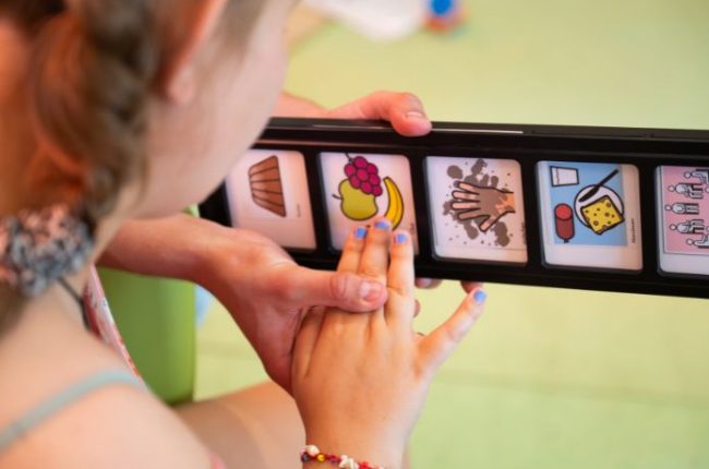 Ein Kind benutzt eine Kommunikationsleiste mit Symbolen, um sich auszudrücken. Die Symbole zeigen verschiedene Bilder wie Essen und Aktivitäten.