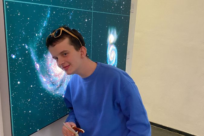 Junger Mann in blauem Sweatshirt steht neben einer Wand auf der eine Galaxie abgebildet ist.