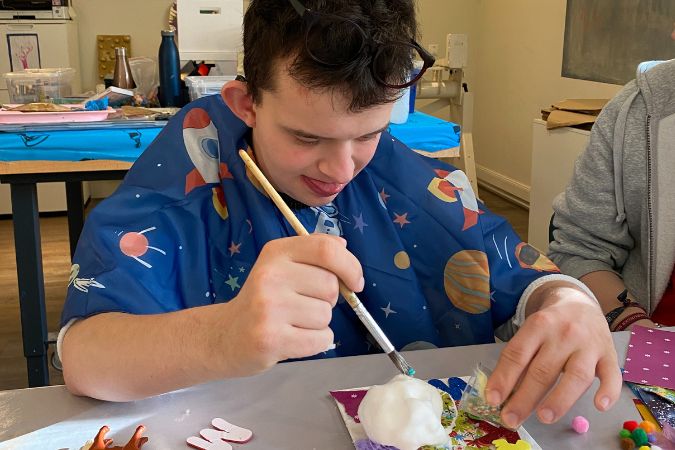 Junger Mann mit Brille, der mit einem Pinsel an einem Kunstprojekt arbeitet, während er einen blauen Umhang trägt.