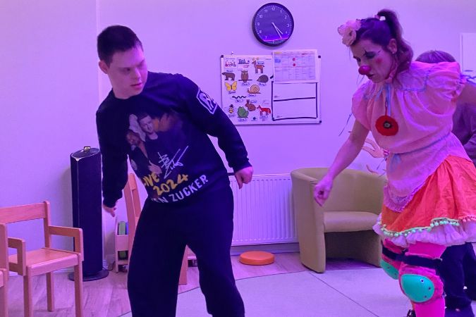 Junge mit Down-Syndrom tanzt mit einer Clownin in einem Raum mit lila Beleuchtung. Die Clownin trägt ein buntes Kostüm und eine rote Nase.