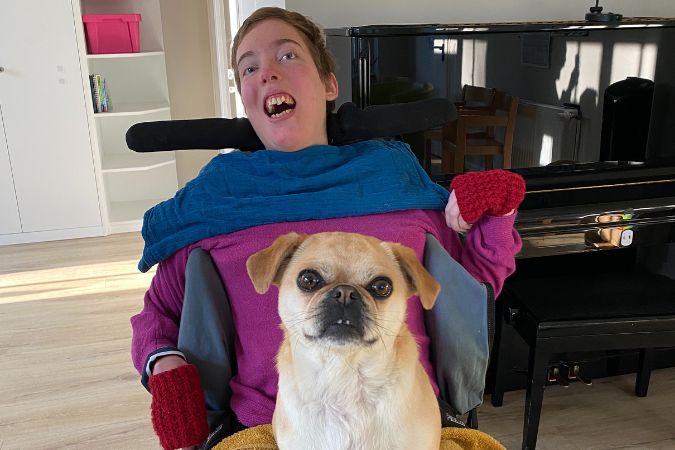 Ein Mädchen in einem Rollstuhl trägt einen blauen Schal und rote Handschuhe, während ein Hund auf dem Schoß sitzt.